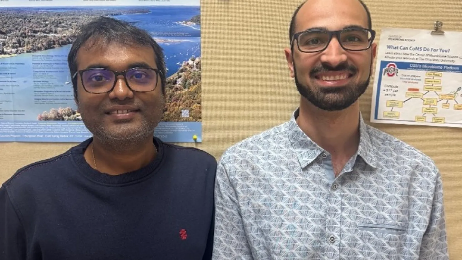 Two men smiling in front of a corkboard
